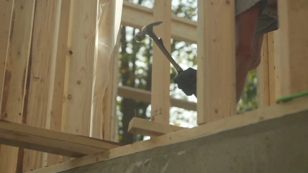 A gloved hand driving a nail into timber stud wall framing on a build site