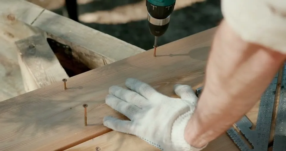 A cordless drill driving a screw into a timber board, steadied by a gloved hand